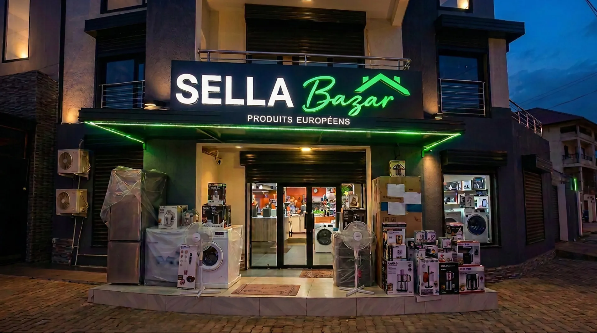 The illuminated Sella Bazar shopfront at night, its sign reading "SELLA Bazar — PRODUITS EUROPÉENS" above a display of washing machines, fans and boxed appliances on the paved forecourt